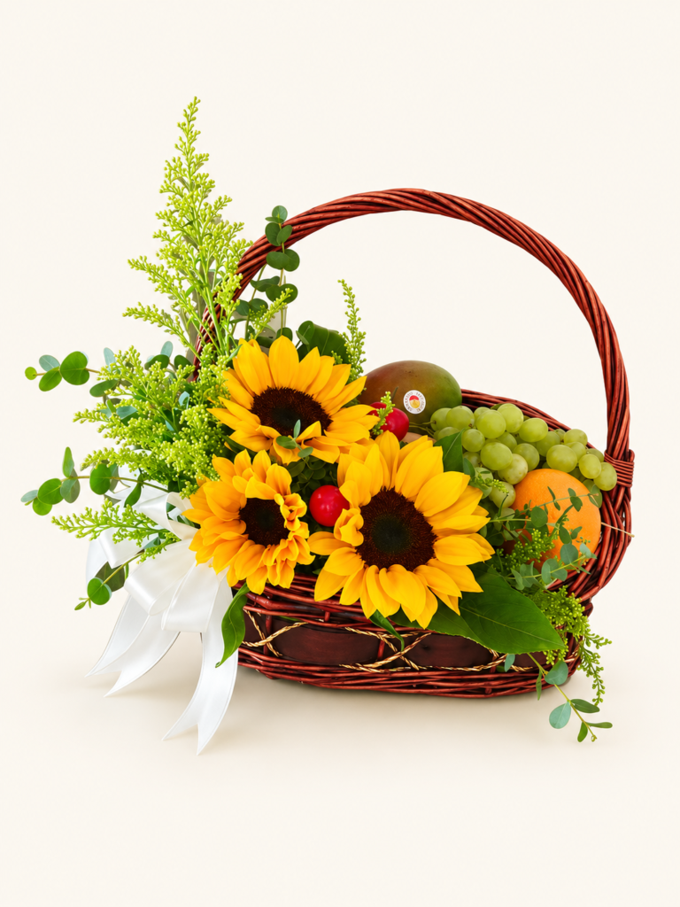 Canasta con asa, girasoles, frutas variadas, follaje y lazo blanco. Sobre fondo crema.