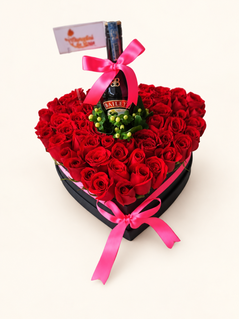Caja en forma de corazón con rosas rojas, botella de Baileys y lazos rosados. Sobre fondo crema.