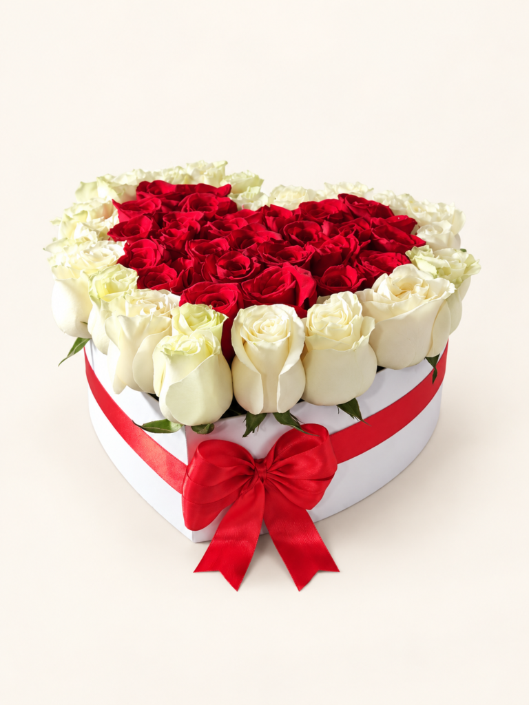 Rosas rojas en el centro y rosas blancas alrededor, en caja corazón blanca con lazo rojo. Sobre fondo crema.